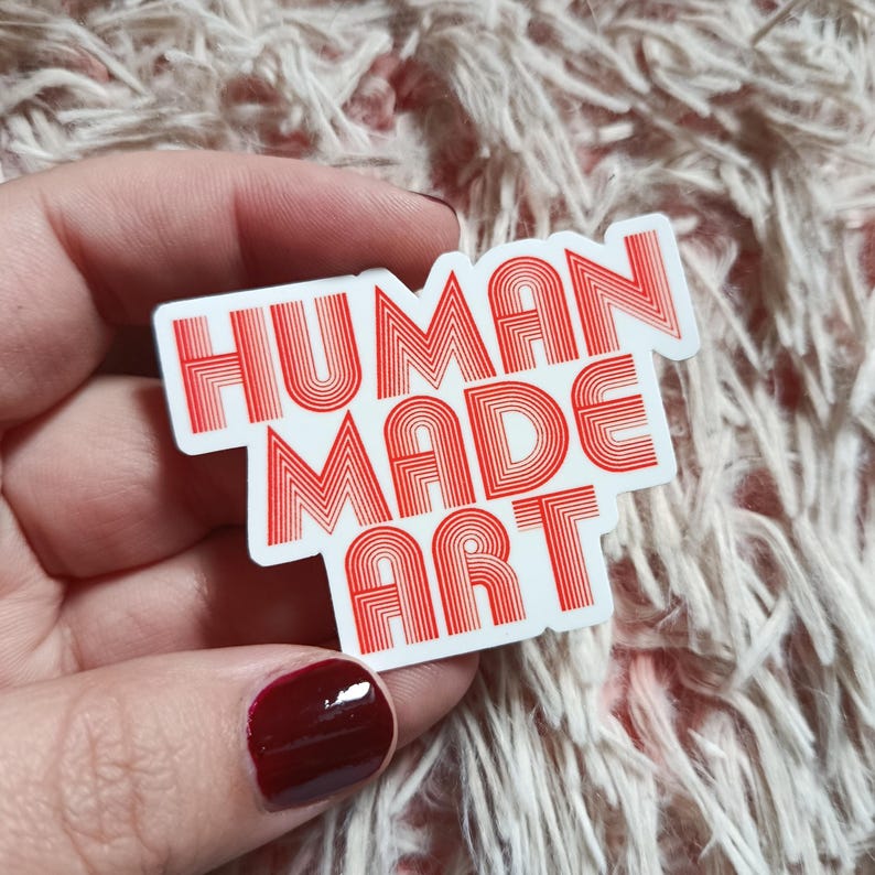 May include: A rectangular sticker with the words "HUMAN MADE ART" in red, retro-style lettering. The sticker is white with a red border and is held by a hand with red nail polish.