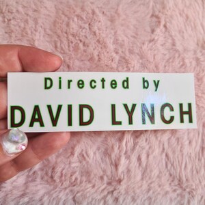 May include: A white rectangular sticker with the text "Directed by DAVID LYNCH" in green and dark red lettering. The sticker is held by a hand with a pink background.