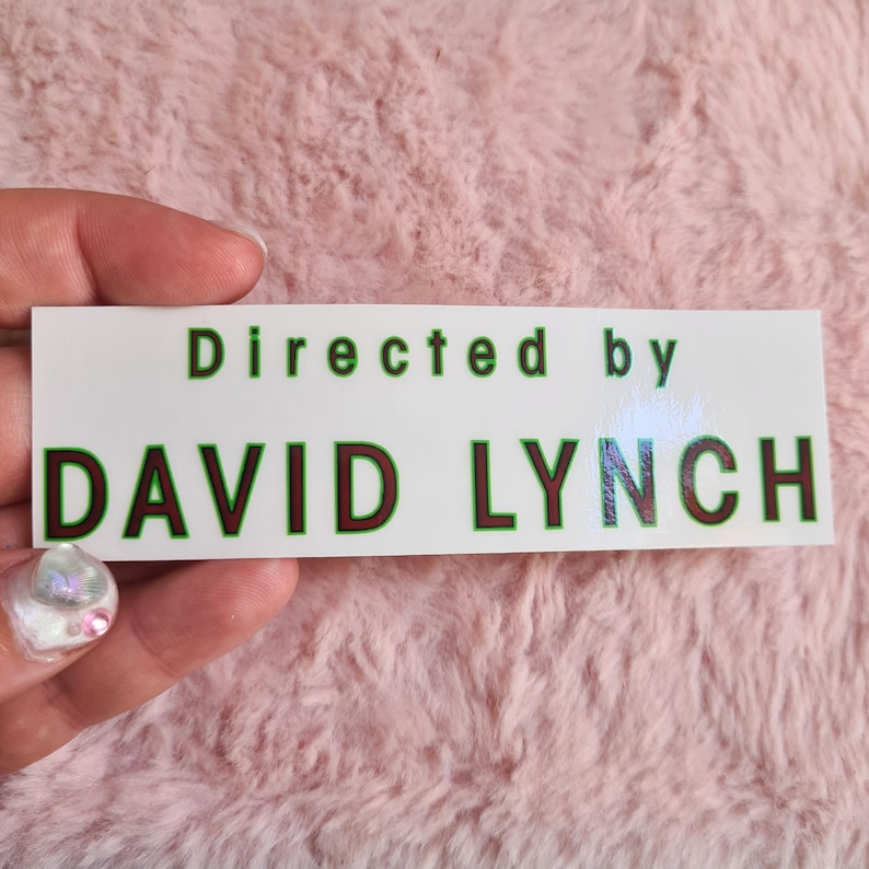 May include: A white rectangular sticker with the text "Directed by DAVID LYNCH" in green and dark red lettering. The sticker is held by a hand with a pink background.