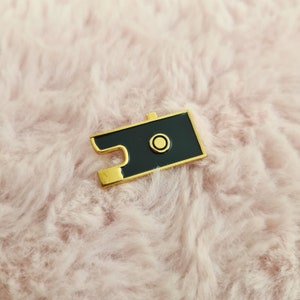 Op de afbeelding: Gouden en zwarte camera emaille pin. De pin heeft de vorm van een zwarte camera met een gouden rand en een gouden cirkel in het midden.