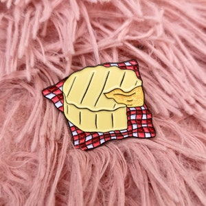 May include: A yellow enamel pin in the shape of a grilled cheese sandwich, with a bite taken out, resting on a red and white checkered napkin. The pin has black outlines and a gold glittery section. The background is a pink, fluffy surface.