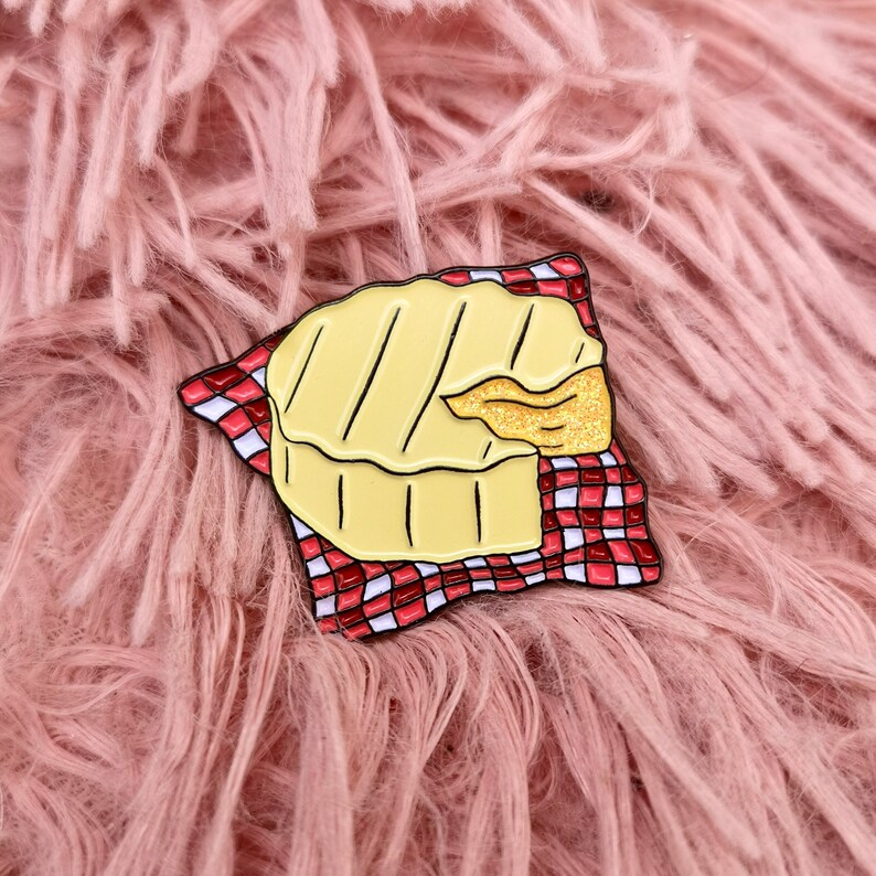 May include: A yellow enamel pin in the shape of a grilled cheese sandwich, with a bite taken out, resting on a red and white checkered napkin. The pin has black outlines and a gold glittery section. The background is a pink, fluffy surface.