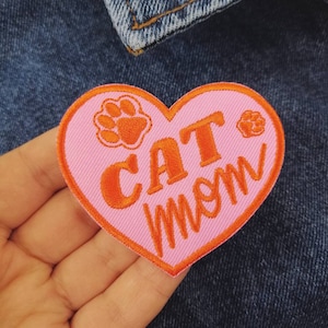 May include: Pink heart-shaped patch with orange embroidered text that reads "CAT MOM" and two paw prints.