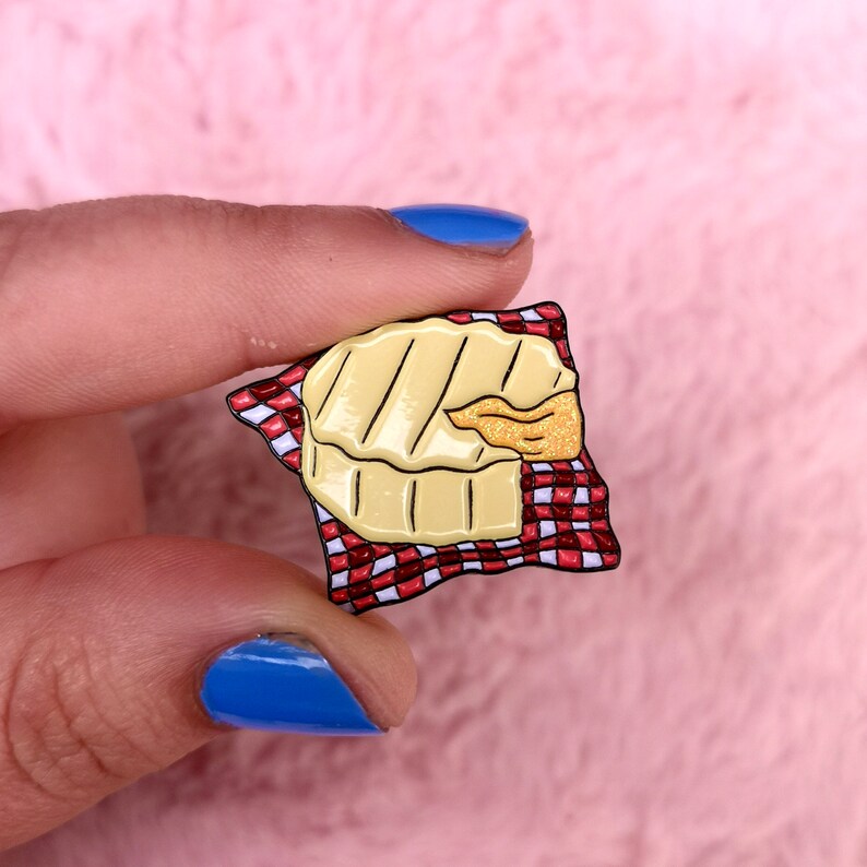 May include: A yellow enamel pin shaped like a grilled cheese sandwich with a golden, glittery cheese pull. The sandwich rests on a red and white checkered napkin. The pin is held against a pink background.
