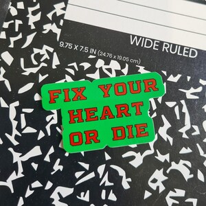 May include: A green sticker with the text "FIX YOUR HEART OR DIE" in red letters. The sticker is on a black and white wide-ruled notebook. The notebook measures 9.75 x 7.5 inches.