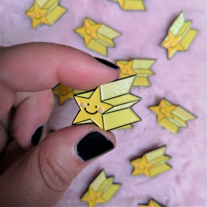 May include: A yellow enamel pin shaped like a shooting star with a smiling face. The star has a glittery trail and is surrounded by other similar pins.