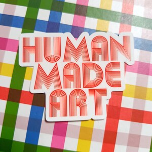 May include: A white sticker with the words "HUMAN MADE ART" in red, retro-style lettering. The sticker is on a colorful plaid background with squares of pink, green, blue, and yellow.