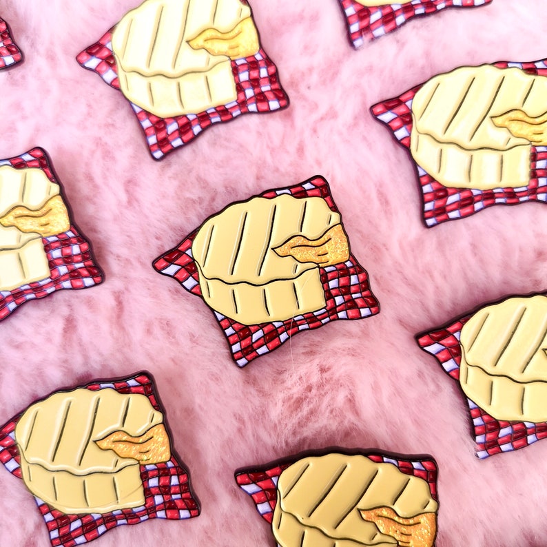 May include: Enamel pins featuring a grilled cheese sandwich on a red and white checkered napkin. The pins have a yellow and gold color scheme, with black outlines. The pins are scattered on a pink, fluffy surface.