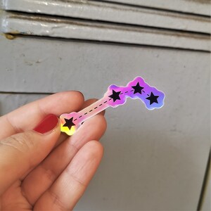 May include: A holographic sticker featuring a constellation of four stars connected by a dashed line. The sticker is iridescent with a pink, purple and blue colour gradient.