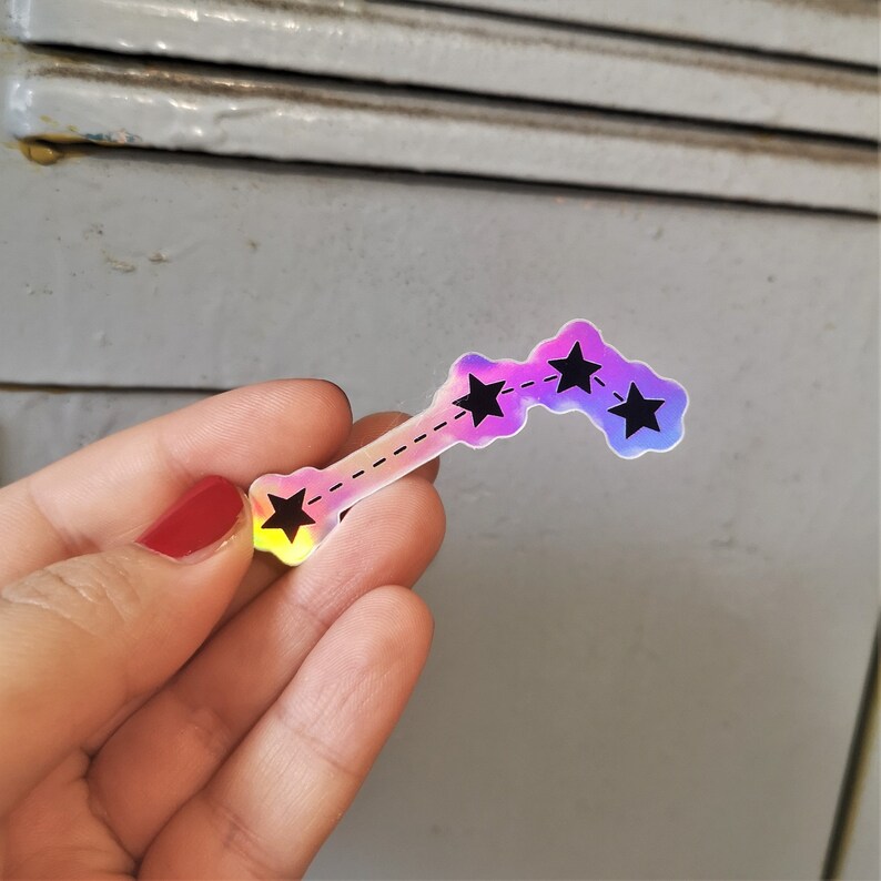May include: A holographic sticker featuring a constellation of four stars connected by a dashed line. The sticker is iridescent with a pink, purple and blue colour gradient.