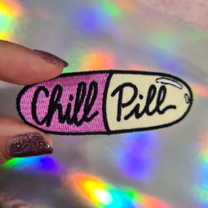 May include: A pink and yellow embroidered iron-on patch shaped like a pill with the text "Chill Pill" in black.