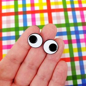 Pode incluir: Dois olhos saltitantes brancos com pupilas pretas, segurados em uma mão contra um fundo xadrez colorido.
