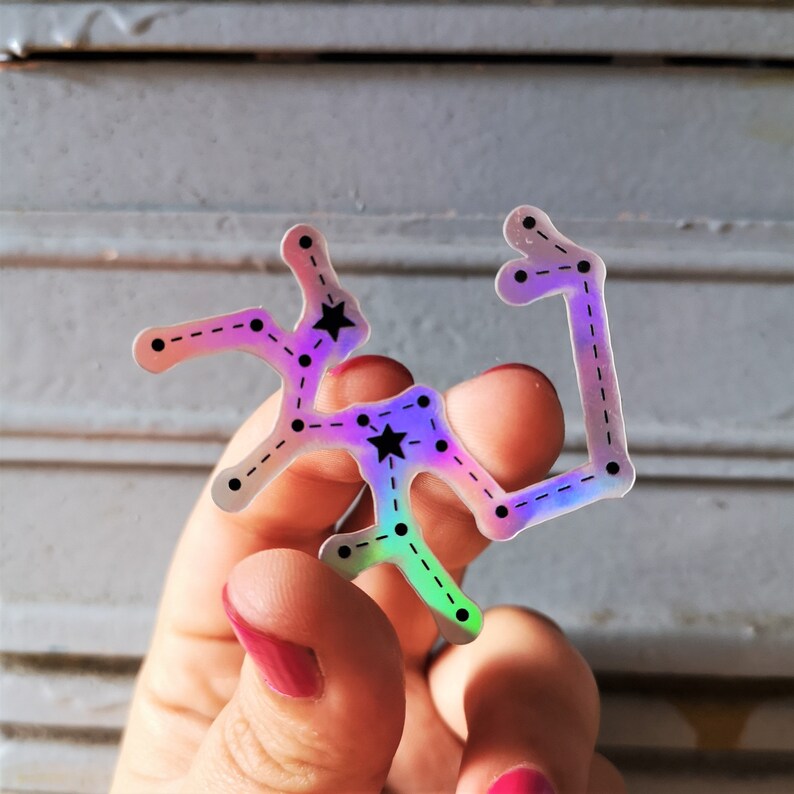 May include: A holographic sticker of a constellation with black stars and a dashed line outline. The sticker is iridescent and has a rainbow effect.
