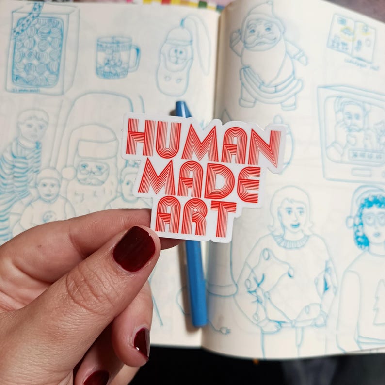 May include: A white sticker with the words "HUMAN MADE ART" in red, layered text. The sticker is held in front of a sketchbook with blue pen sketches of various objects and figures.