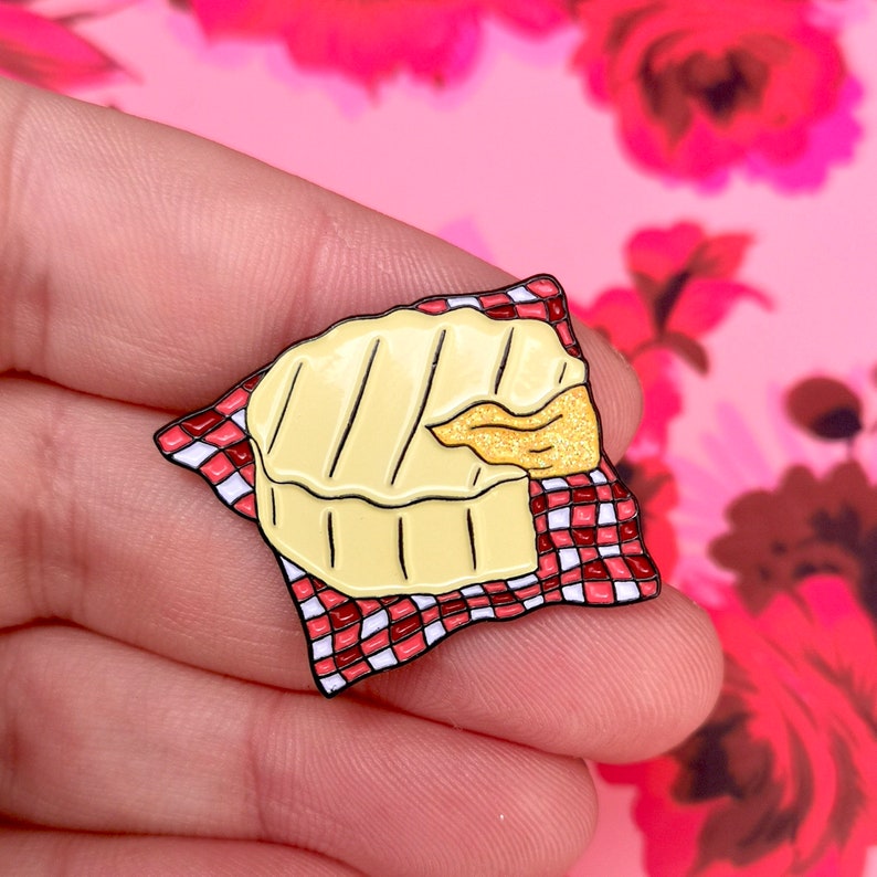 May include: A cheese enamel pin featuring a wheel of brie with grill marks, resting on a red and white checkered napkin. A golden, glittery section of cheese is visible. The pin is set against a pink floral background.