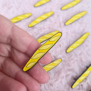 Può includere: Uno spillo in smalto giallo con glitter a forma di baguette. Lo spillo ha un contorno nero ed è tenuto in una mano.