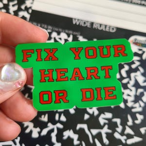 May include: A green sticker with the words "FIX YOUR HEART OR DIE" in red block letters. The sticker is held in a person's hand, with a notebook visible in the background.