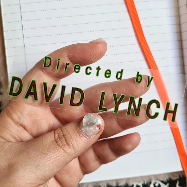 May include: A clear, rectangular sticker with the text "Directed by DAVID LYNCH" in green letters with a red outline. The sticker is held over a notebook with lined paper and an orange ribbon.