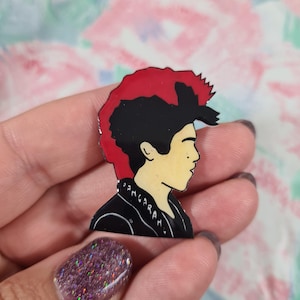 May include: A black and red enamel pin depicting a profile of a person with a mohawk hairstyle. The person is wearing a black leather jacket with the word "Ramones" written on it.