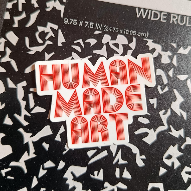 May include: A white sticker with the words "HUMAN MADE ART" in red, retro-style lettering. The sticker is on a black and white patterned background. The notebook measures 9.75 x 7.5 inches.