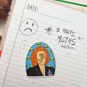 Può includere: Un adesivo con un'illustrazione di Dana Scully dei X-Files. Scully indossa una giacca di un completo nero con un distintivo dell'FBI e ha i capelli rossi. Lo sfondo è blu con teste di alieni verdi.