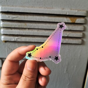 May include: A holographic sticker featuring a constellation of stars in a rainbow colour scheme. The sticker is shaped like a triangle and has a dotted line connecting the stars.