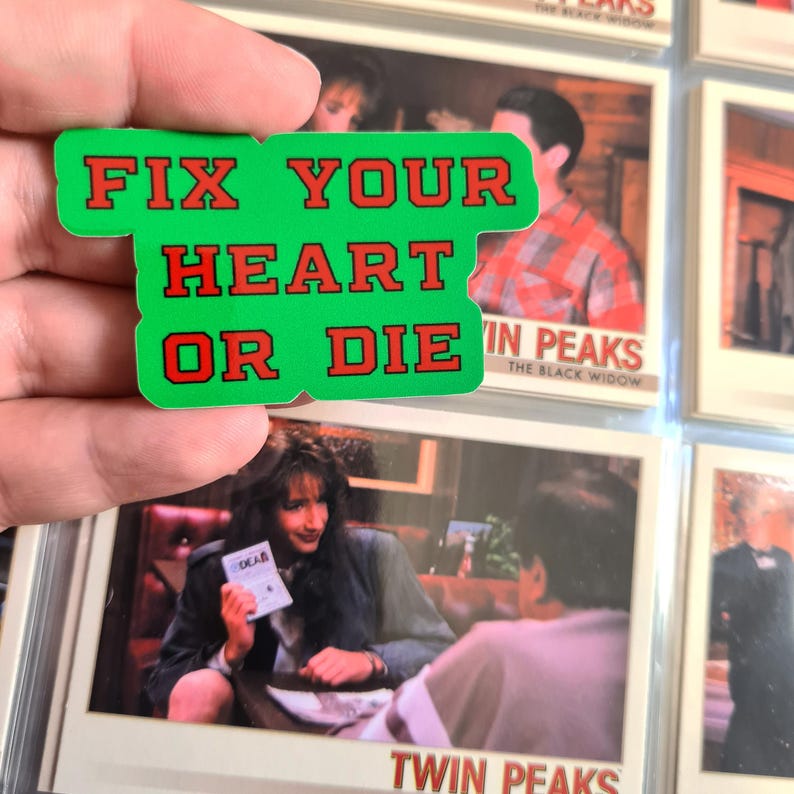 May include: A green rectangular pin with the words "FIX YOUR HEART OR DIE" in red lettering. The pin is held in front of a collection of Twin Peaks trading cards.