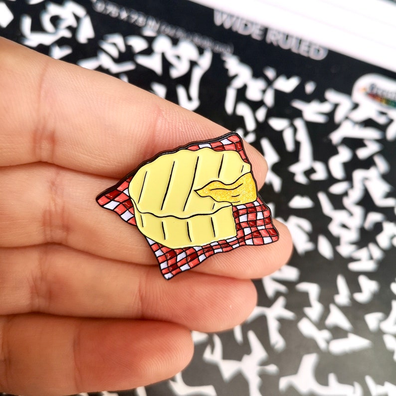 May include: A yellow enamel pin depicting a piece of brie cheese on a red and white checkered napkin. The pin has black outlines and is held in a hand. The cheese has a golden, melted section.