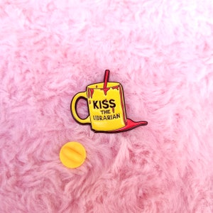 May include: Yellow enamel pin shaped like a coffee mug with a red straw and red dripping down the side. The mug says "Kiss the Librarian".