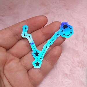 May include: A holographic blue sticker with a black outline of the Pisces constellation. The constellation is made up of stars and dots.