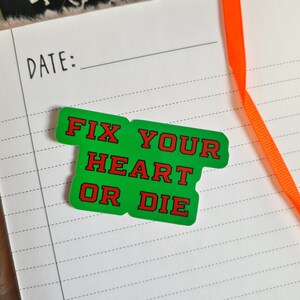 May include: A green sticker with red text that reads "FIX YOUR HEART OR DIE" is placed on a lined notebook page. An orange ribbon is visible in the background. The word "DATE:" is printed at the top of the page.