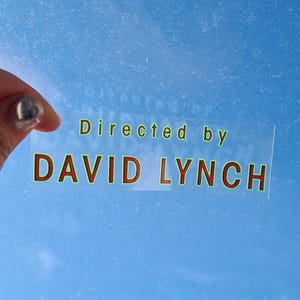 May include: A clear sticker with the text "Directed by DAVID LYNCH" in green and red lettering. The sticker is held against a light blue background.