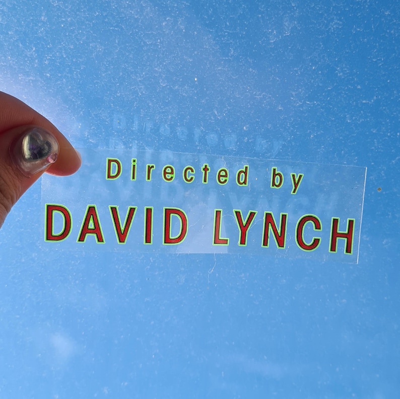May include: A clear sticker with the text "Directed by DAVID LYNCH" in green and red lettering. The sticker is held against a light blue background.