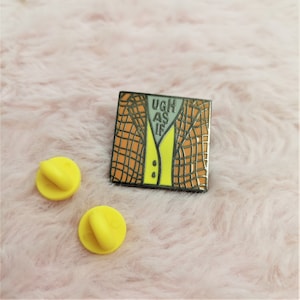 May include: A yellow and orange enamel pin with a cartoon image of a person wearing a plaid jacket and the text "UGH AS IF".