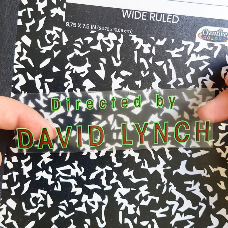 May include: A notebook with a black and white pattern. The notebook is labeled "WIDE RULED" and measures 9.75 x 7.5 inches. A clear sticker with the text "Directed by DAVID LYNCH" is placed over the notebook.