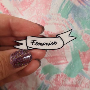 Può includere: Uno spillo in smalto bianco con un contorno nero a forma di striscione. Lo striscione recita "Femminista" in corsivo nero.