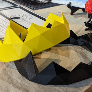 May include: A yellow and black 3D printed crown. The crown is made of geometric shapes and has a pointed design.