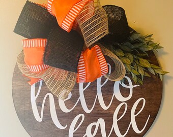 May include: A round wooden sign with the words "hello fall" in white script. The sign is decorated with orange and black ribbons, burlap, and greenery, perfect for autumn decor.