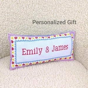 May include: A rectangular, white cross-stitch pillow with the names "Emily & James" in red. The pillow has a decorative border with red, yellow, and blue accents. The text "Personalized Gift" is above the pillow.