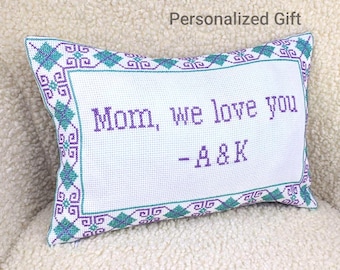 May include: A white rectangular pillow with a cross-stitched design. The pillow features the text "Mom, we love you - A&K" in purple. The border is decorated with teal and purple geometric patterns. The words "Personalized Gift" are above the pillow.