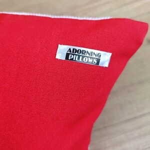 Detail shot of the fabric label on a square red black personalized decorative pillow cover, showing the 'Adorning Pillows' logo as proof of artisan quality and a unique handmade brand.