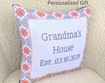May include: A white cross-stitch pillow with the words "Grandma's House" and "Est. 03.18.2026" in blue. The pillow has a decorative border in coral and blue, with the text "Personalized Gift" at the top.
