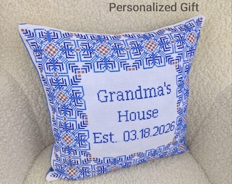 May include: A square throw pillow featuring a white center with blue cross-stitched text reading Grandma's House Est. 03.18.2026. The border displays a geometric pattern in shades of blue and brown, creating a decorative frame around the text.
