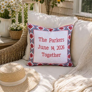 May include: White square pillow with red and blue embroidered border. The pillow features the text "The Parkers June 14, 2026 Together" in red. A straw hat rests below the pillow on a white cushion.