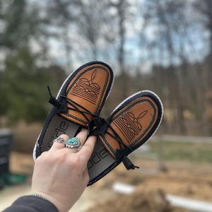 Puede incluir: Un par de zapatos de cuero negro con detalles en cuero marrón y costuras turquesa. Los zapatos tienen cierre de cordones y suela blanca. La palabra "Dude" está impresa en el interior del zapato.