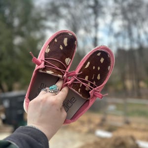May include: A pair of pink casual shoes with a brown and white animal print lining. The shoes are held in a hand with a ring on the finger. The brand name "JUCY" is visible on the insole.