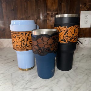 May include: Three insulated tumblers with leather wraps. The tumblers are blue, black, and light blue. The leather wraps have floral and leaf designs.
