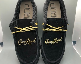 May include: A pair of black slip-on shoes with a gold Crown Royal logo embroidered on the front. The shoes have a black mesh upper and a brown leather insole.