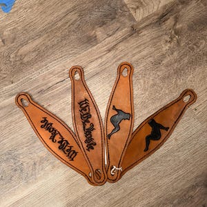 Puede incluir: Cuatro pulseras de cuero marrón con diferentes diseños. Una pulsera tiene el texto "We Be Trouble" en letras negras. Otra pulsera tiene el texto "1028" en letras negras. Las otras dos pulseras tienen una silueta de una mujer en negro.