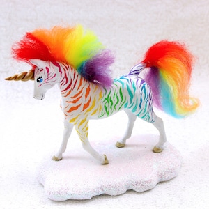 May include: A whimsical unicorn figurine with a rainbow mane and tail. The unicorn has a white body with colorful zebra stripes. It stands on a glittery white cloud base. The horn is a golden brown color.